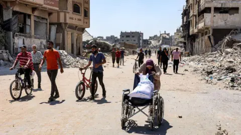 AFP A woman pushes a wheelchair loaded with a sack of flour as she walks past rubble and damaged buildings along a street in the Tuffah district of Gaza City, in northern Gaza (8 July 2024)