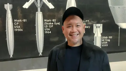 Photograph of Low Kee Hong from Factory International. he is pictured in front of a mural made of Lego bricks at Manchester's Aviva Studios.