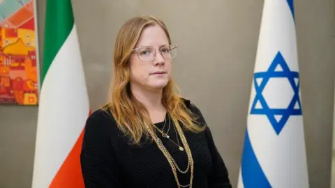 PA Media Erlich is standing in between the blue and white Israeli flag and the green white and orange, Irish tricolour flag. She has long blond hair and is wearing thin framed glasses and a black top. She is also wearing a gold necklace.