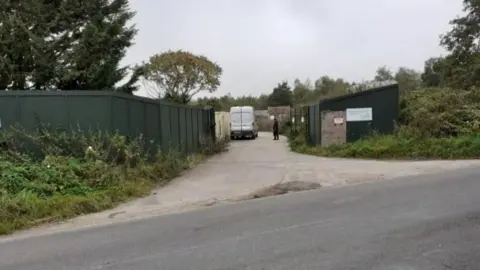 West Berkshire Council Dark green hoarding is either side of the entrance which is approached on a concrete driveway. A white van can be seen just inside the entrance and a person wearing dark clothing standing near it inside the entrance gates