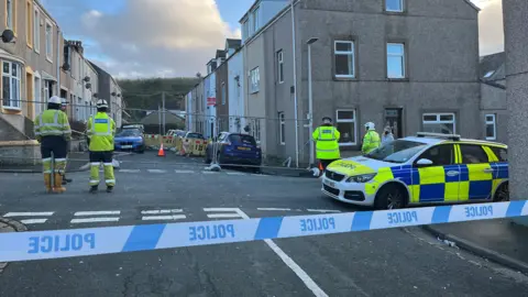 Officers in high-vis jackets stand on a street, which is partly fenced off. A police car and police tape are in the foreground.