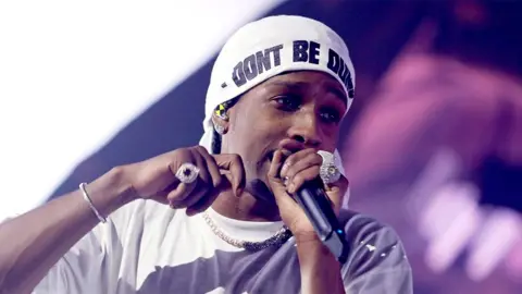 Getty Images A$AP Rocky performs on stage singing into a black microphone while wearing a white t-shirt and a white cap with the inscription "Don't be stupid"silver chain around her neck and sparkling rings on her fingers.