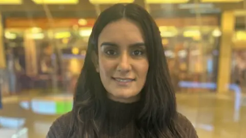 BBC Kiranjit is stood outside the BBC newsroom in Birmingham. She has long dark hair and a brown top on and is smiling at the camera. The newsroom is blurred in the background.
