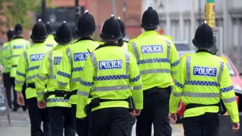 A group of Humberside Police officers marching down a street in Hull. They have their backs to the camera.  They are wearing fluorescent jackets with the word "police" on the back. They are all wearing traditional black and silver police helmets