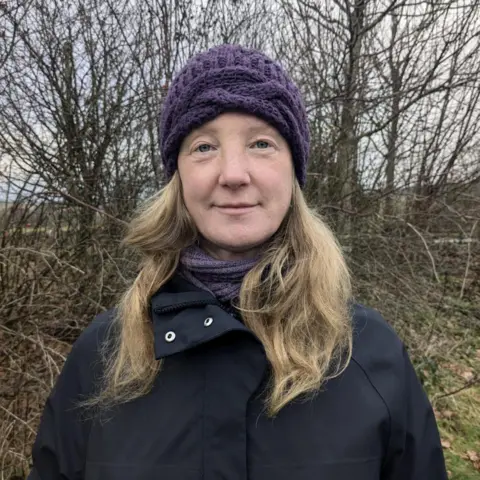 Woman smiling at the camera with bare trees behind her. She has long, blonde hair and is wearing a black jacket, purple scarf and purple hat.