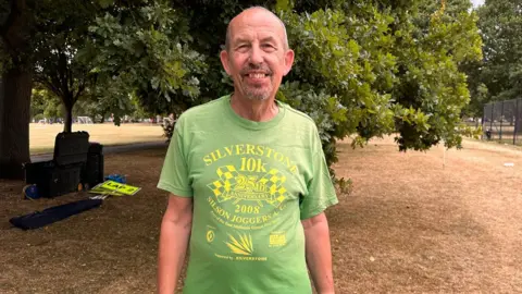 Carroll Weston/BBC Bob Emmerson (junior) who has short grey hair and a salt and pepper goatee beard. He is wearing a faded green T-Shirt which has Silverston 10k 25th anniversary 2008 written on it in yellow letters. Behind him is a large tree and parched-looking yellow grass.