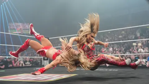 Getty Images Two women are falling to the ground in a wrestling ring. They are both wearing red with long blonde hair. 