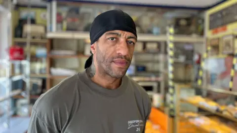 George Carden/BBC Gareth Lowndes who has a beard, wearing a durag and has a tattoo on his neck standing in the bakery with pastries behind him