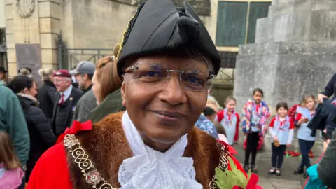 Bharat Pankhania. He is dressed in mayoral attire which includes a red robe and a black hat. He is looking at the camera and smiling.