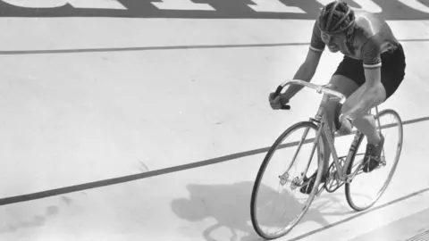 A black-and-white image shows a cyclist riding a track bike on a velodrome.

The cyclist is leaning forward in a racing position, pedalling along a marked lane.