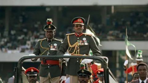 Sygma via Getty Images Buhari, wearing a military uniform, stands at the back of an open top vehicle. Three other men in military gear are also in the car, which appears to be in an arena.
