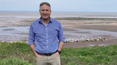 Councillor Mike Rigby wearing light grey trousers and a blue and white chequered shirt. He is standing with his hands in his front pockets on a grassy patch beside a beach.
