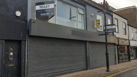 The site which has been given approval. A disused commercial front with black shutters, with an upper story with windows facing outwards. Next door are other businesses on the street. "To let" signs are posted outside the space.
