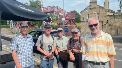 Frank Phillips, Peter Etherington, Rob Doughty, Tony Evans and Gerry Slater standing it front of a railway bridge in the sun. They are all smiling in a beer garden wearing short-sleeve tops.