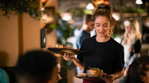 Getty Images Waitress wearing a black T-shirt, burgundy apron and high ponytail serves two plates of Mexican food to restaurant diners. 