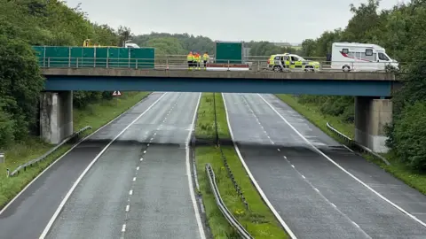 BBC The empty A1 which is a dual carriageway with a central reservation. There is a bridge above the road with emergency service workers in hi-vis standing on it looking down. There is also a police car and caravan parked.