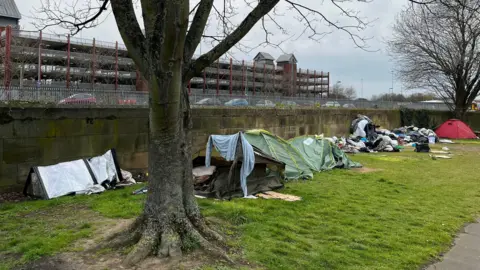 A row of tents have been erected in front of a wall on a grassy strip of land. A pile of mattresses and rubbish can be seen further down the wall.