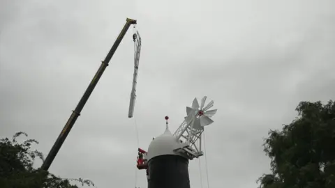 A crane lifts a white wooden windmill sail to the top of Skidby Windmill. The building is black with a white domed top with a smaller windmill on it 
