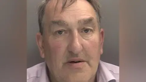 A mugshot of a man with dark grey hair. He wears a light pink shirt.