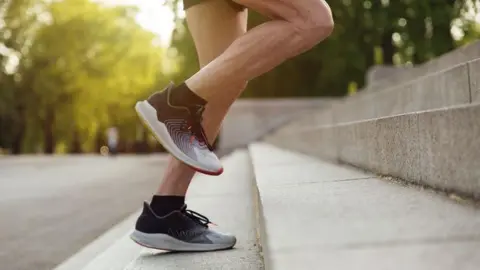 A man's legs whilst they're running outdoors. The person is running up a set of stone stairs and they're wearing running shoes and black socks.