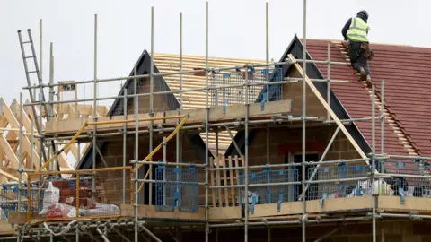 A builder on the roof of a partially built house with scaffolding in the front.