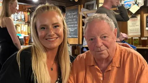 Contributed Beth and Lawrence Dixon are sitting next to each other in a pub. Beth is on the left and has long blonde hair and is wearing a black cardigan and a black and white top. Lawrence is on the right and is wearing an orange shit and glasses are resting on top of his head.