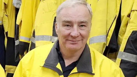 RNLI/Tom Collins Mark Barker. He is wearing his bright yellow RNLI coat. He has grey hair and is smiling at the camera. Behind him a number of yellow jackets are hung on hooks in a row.