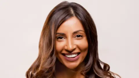 Gtech A woman with long brown hair is prominent in the photo and smiling at the camera in front of a plain light background.