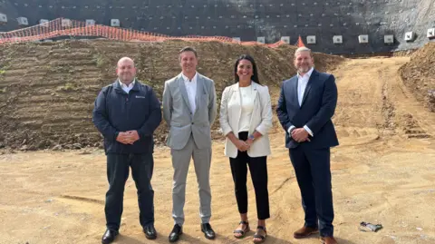 A picture of four people stood in a line at the site where the proposed homes are set to be built. It is a large open space.