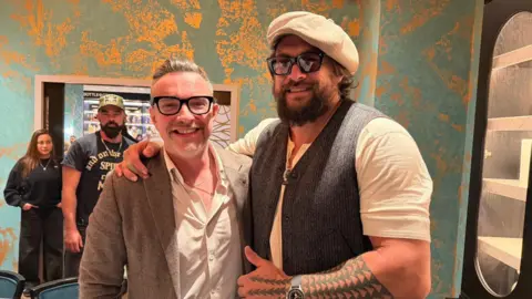 Ed James and Momoa stand together in a smartly decorated room. James has glasses on and a beige suite and shirt. Momoa, in a white T-shirt and grey waistcoat and flat cap, has his arm around him.