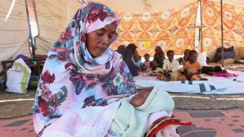 Reuters A woman in a headscarf, sitting on the floor in a large tent cradling a baby.