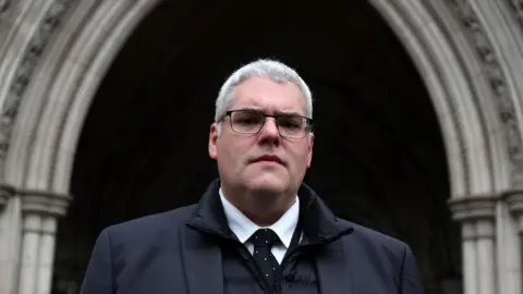 EPA A man with white hair and black glasses is standing outside an ornate building. He is wearing a black suit over a black coat white shirt and tie.