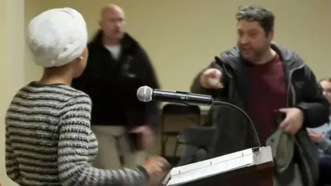 Man points at Ilhan Omar, who is standing behind a lectern.