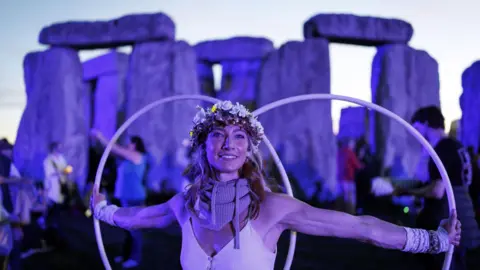 PA Media A woman wearing a white top and a floral headdress stands near the stones of Stonehenge just before dawn. She has long wavy blonde hair and is holding a white hula hoop with each hand