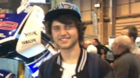 Nadine Marshall Conner, wearing a cap and a baseball jacket, smiles as he stands next to a motorbike