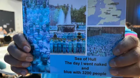 A close up of a man holding a blue book it reads 'Sea of Hull the day I went naked and blue with 3200 people'