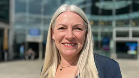 Kay Mason Billig smiling and wearing a dark blue suit jacket. It is clearly sunny weather. She is standing in front of The Forum in Norwich, a glass-fronted building.