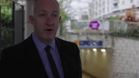 Ray Morgon who is wearing a dark blue suit, a lighter blue shirt and a blue tie, standing in front of an underpass. 