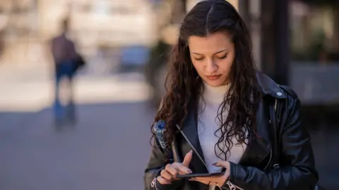 Getty Images A young woman wearing a leather jacket over a white jumper is standing in a street checking her smartphone.
