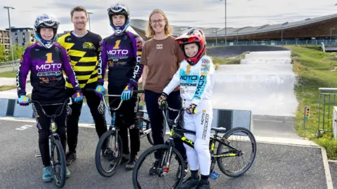 Better.org.uk The family pose for a photo, with the children still holding their bikes. Andy and Alex stand with them smiling. The track and seating area can be seen in the background. 
