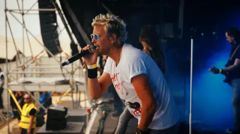 Man with blonde hair and translucent framed sunglasses with blue lenses. He's wearing a white t shirt and singing into a microphone