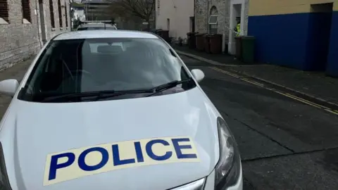 A police car parked in Rendle Street with a scene guard standing in the door way of a house behind police incident tape. 