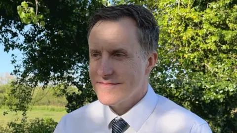 A man wearing a white shirt and a blue stripy tie standing in front of some greenery.