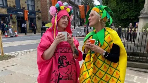 University of Leeds Two people dressed up in fancy dress. One is dressed as a pink Whoopie cushion, and one is dressed as a pineapple. They stand on a residential street holding mugs of tea.