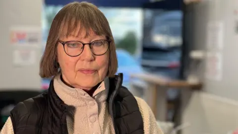 BBC/Gill Dummigan Annabel Marsh has bobbed brown hair and is wearing glasses. She is wearing a cream fleece top and a black padded gilet and standing in a room. A TV screen can be seen blurred out behind her.