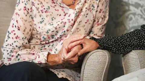 PA MEDIA A hand clasped around the arm of an elderly woman. both are leaning on the arms of grey armchairs. The arm of the person on the right is clad in the sleeve of a black blouse with small white polka dots. The elderly lady is wearing a light coloured blouse with a a green, yellow and pink floral pattern and dark trousers. She is also wearing a beaded necklace.