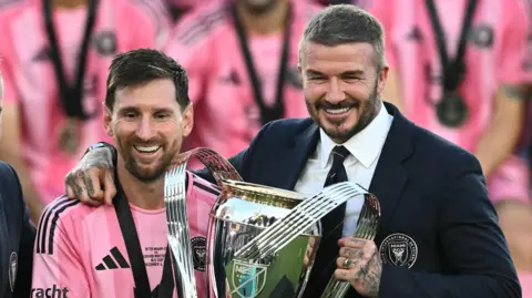 Lionel Messi stands alongside David Beckham, who has his right arm around the Argentine as they show off the MLS Cup trophy. Messi is wearing Inter Miami's pink kit, while Beckham is dressed smartly in a club blazer, white shirt and dark tie. Both are smiling.