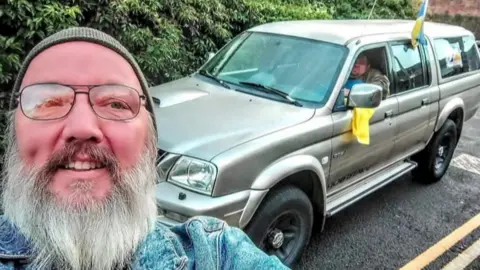 Pete Laschok Pete stood in front of a 4x4 vehicle with a Ukrainian flag on top of it. Pete is a man with a grey beard, glasses and a beanie hat on his head.