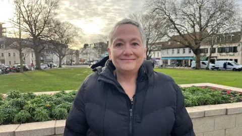 The photo shows a woman standing outside, with short grey hair and wearing a black coat. She is standing in front of a flowerbed, with grass and trees behind. Beyond those are shops.