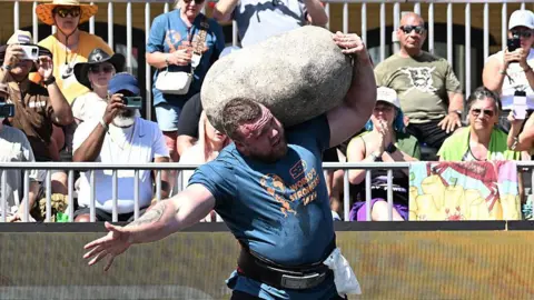 Getty Images Tom Stoltman competing in World's Strongest Man. She is holding a large stone on his shoulder. 